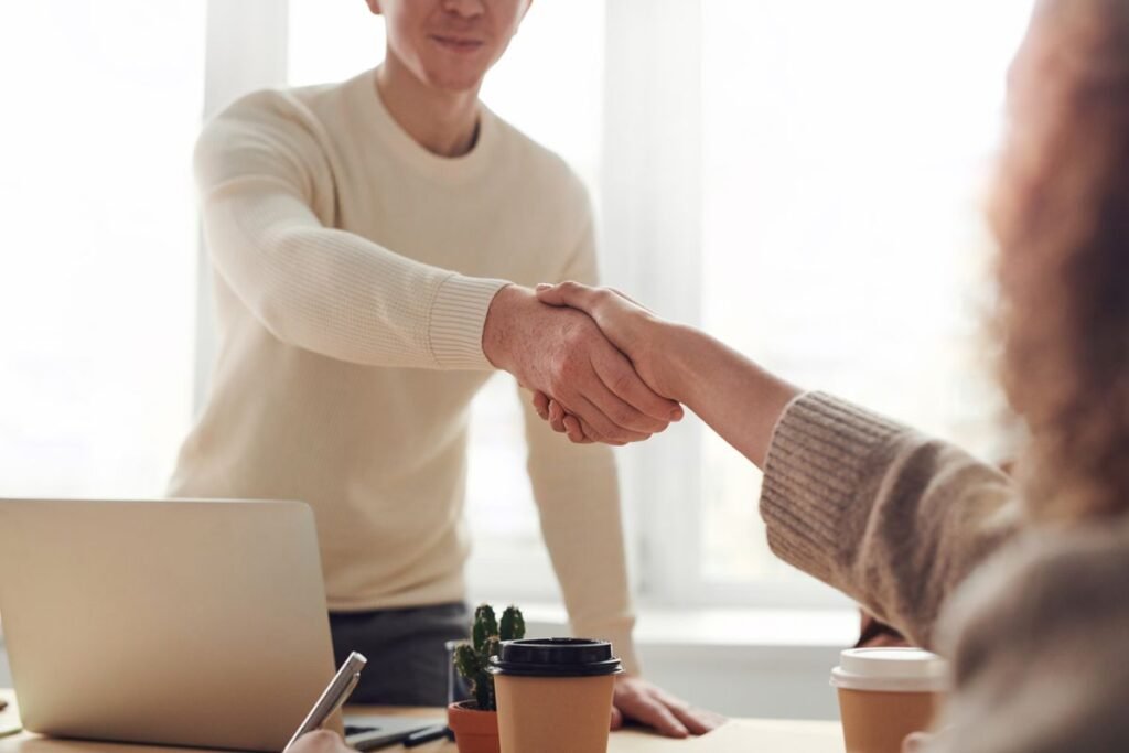 Two business people shaking hands over a desk with laptop and coffee
